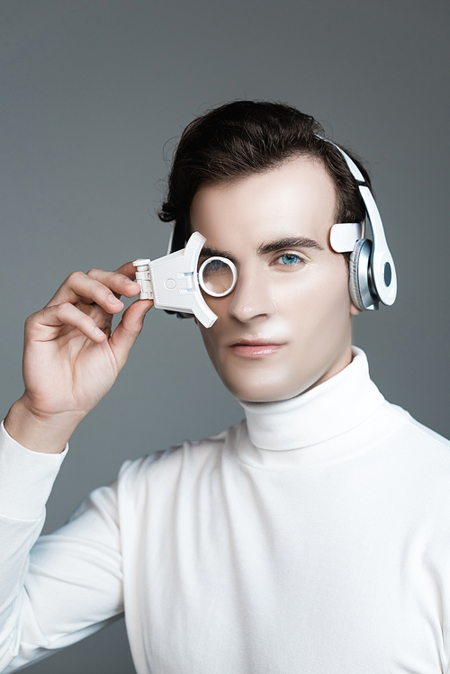 Brunette cyborg in headphones holding digital eye lens isolated on grey