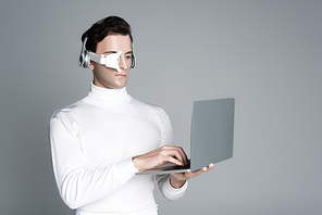 Cyborg in eye lens and headphones using laptop isolated on grey