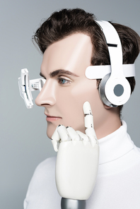 Cyborg man with headphones and finger of artificial hand near cheek isolated on grey