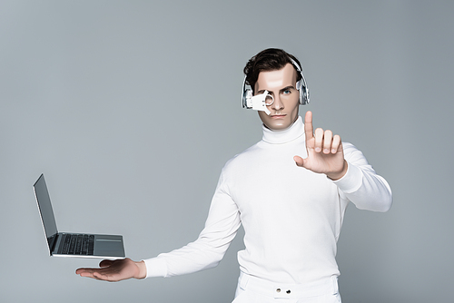 Cyborg in headphones touching something near laptop with blank screen levitating in air isolated on grey