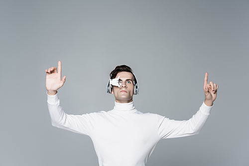 Brunette cyborg man in digital eye lens and headphones looking away while using something isolated on grey