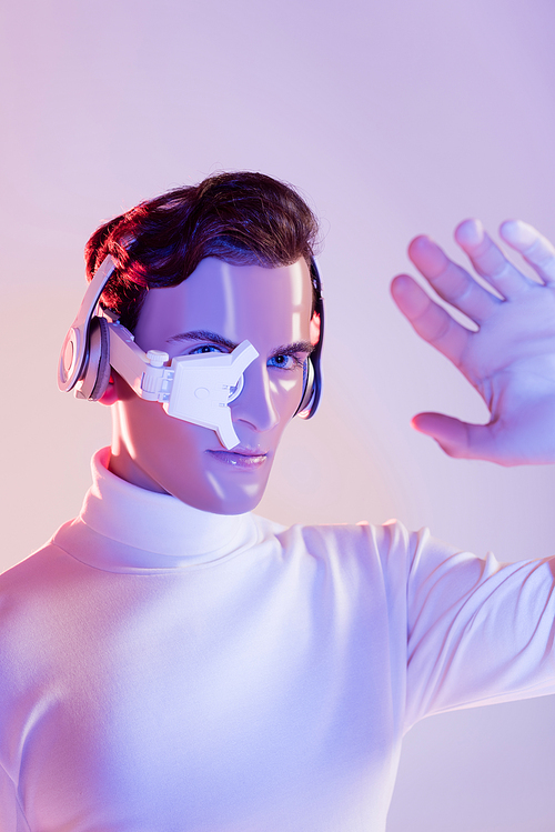 Cyborg man in eye lens and headphones looking at hand on blurred foreground on purple background