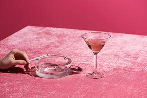 cropped view of woman with cigarette near ashtray and glass of rose wine on velour pink cloth isolated on pink, girlish concept