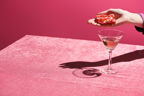 cropped view of woman holding rose petals above glass of rose wine on velour cloth isolated on pink, girlish concept
