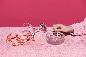 cropped view of woman putting out cigarette near glass with scattered petals on velour cloth isolated on pink, girlish concept