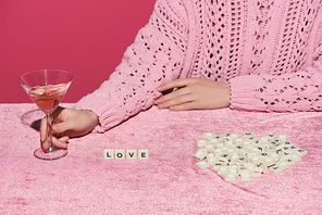 cropped view of woman holding glass of rose wine on velour cloth with love lettering on cubes isolated on pink, girlish concept