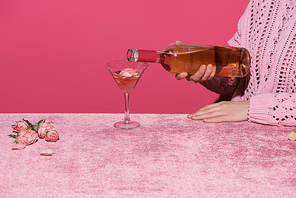 cropped view of woman pouring rose wine near rose petals on velour cloth isolated on pink, girlish concept