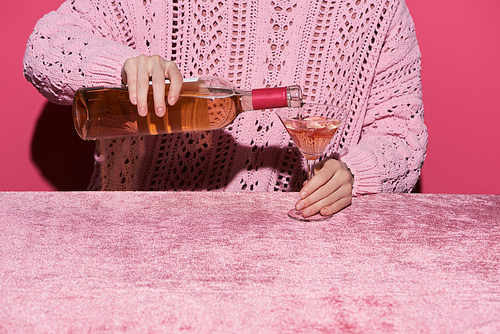 cropped view of woman pouring rose wine from bottle into glass on velour cloth isolated on pink, girlish concept