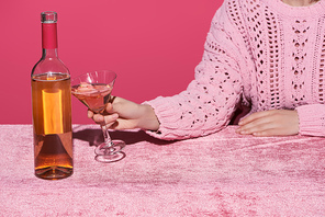 cropped view of woman holding glass of rose wine near bottle on velour cloth isolated on pink, girlish concept