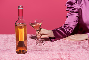 cropped view of woman holding glass of rose wine near bottle on velour cloth isolated on pink, girlish concept