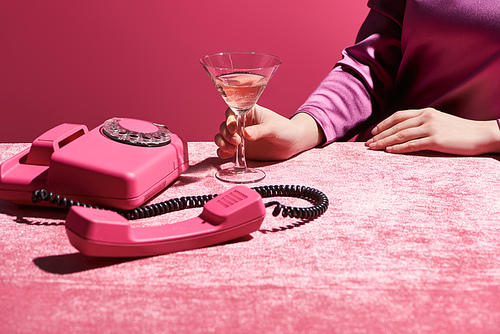 cropped view of woman holding glass of rose wine near retro phone on velour cloth isolated on pink, girlish concept