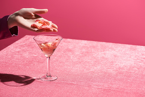 cropped view of woman holding petals above glass on velour cloth isolated on pink, girlish concept