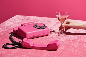 cropped view of woman holding glass of rose wine near retro phone on velour cloth isolated on pink, girlish concept