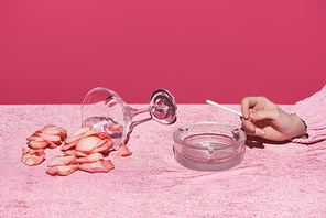 cropped view of woman with cigarette and ashtray near glass with rose petals on velour cloth isolated on pink, girlish concept