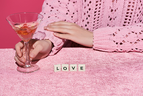 cropped view of woman holding glass of rose wine near cubes with love lettering on velour cloth isolated on pink, girlish concept