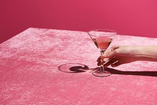 cropped view of woman holding glass with drink on velour cloth isolated on pink, girlish concept