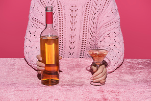 cropped view of woman holding bottle of alcohol drink and glass at velour pink table isolated on pink, girlish concept