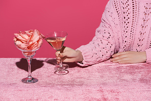 cropped view of woman holding glass of rose wine near glass with rose petals on velour cloth isolated on pink, girlish concept