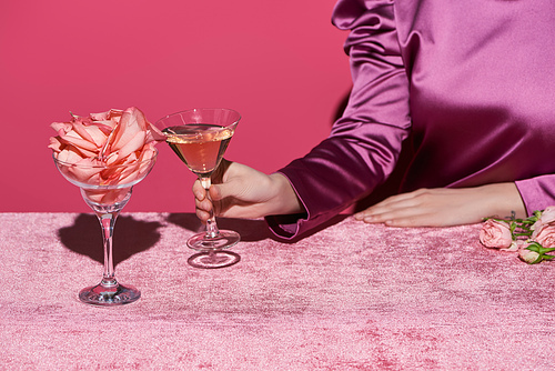cropped view of woman holding glass of rose wine near glass with rose petals on velour cloth isolated on pink, girlish concept
