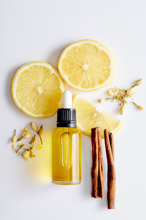 Top view of bottle of cosmetic oil with slices of lemon, sticks of cinnamon and vanilla buds on white background