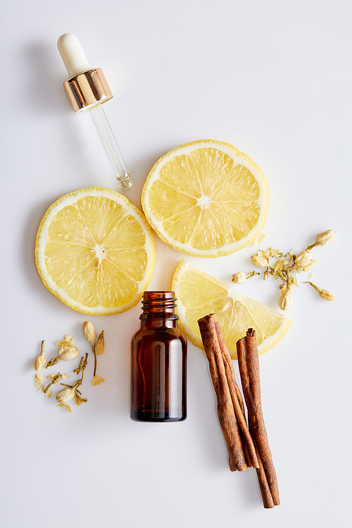 Top view of bottle of cosmetic oil with dropper, slices of lemon, sticks of cinnamon and vanilla buds on white background
