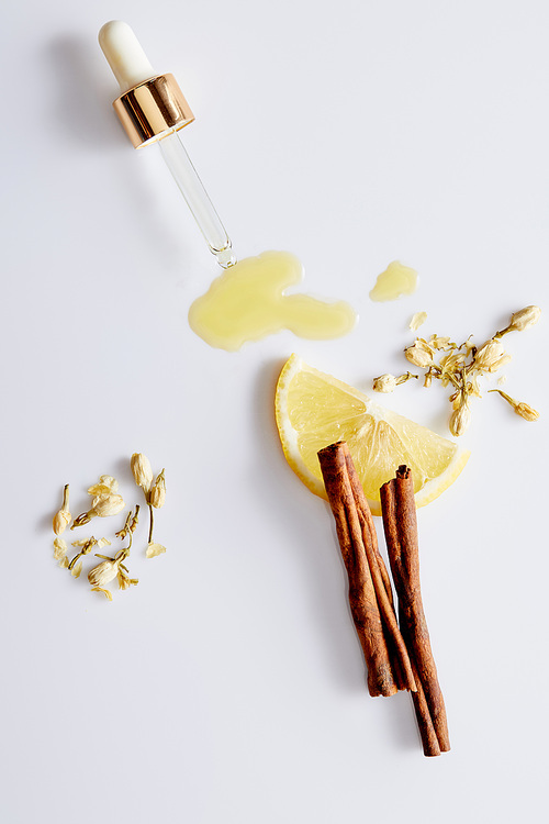 Top view of cosmetic oil flowing out of dropper next to slice of lemon, sticks of cinnamon and vanilla buds on white background