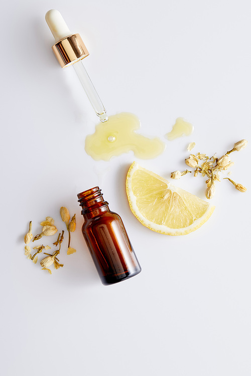 Top view of cosmetic oil flowing out of dropper next to bottle, slice of lemon and vanilla buds on white background