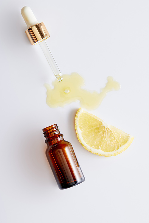 Top view of cosmetic oil flowing out of dropper next to bottle and slice of lemon on white background