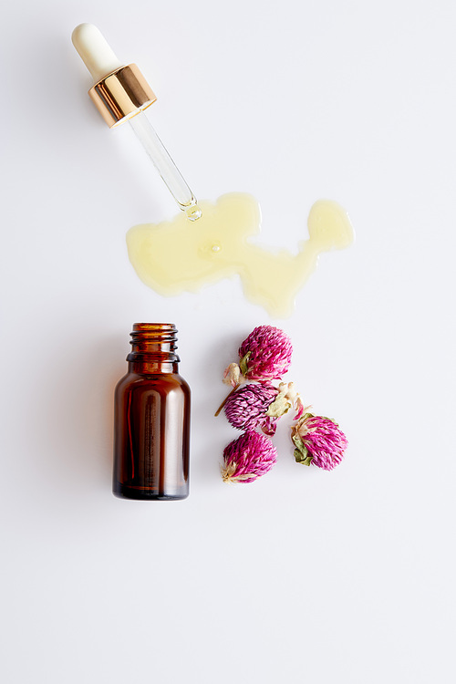Top view of cosmetic oil flowing out of dropper next to bottle and pink buds on white background