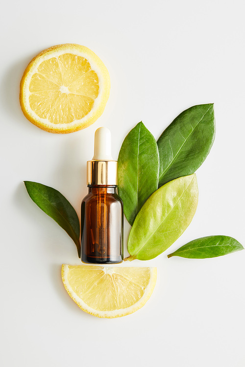 Top view of bottle of cosmetic oil with leaves and slices of lemon around on white background