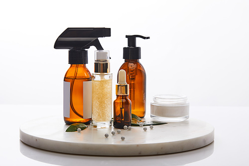 Dispenser bottles of oil and cosmetic cream next to leaves, decorative beads on round stand isolated on white