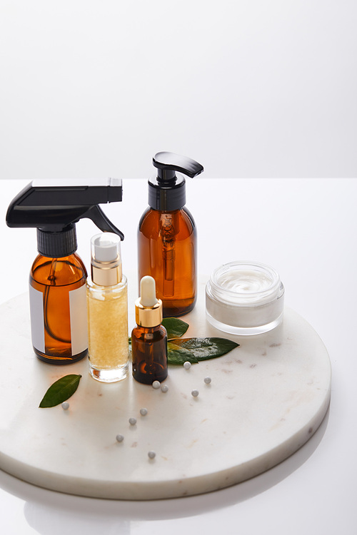 Dispenser bottles of oil and cosmetic cream with leaves and decorative beads on round stand isolated on grey