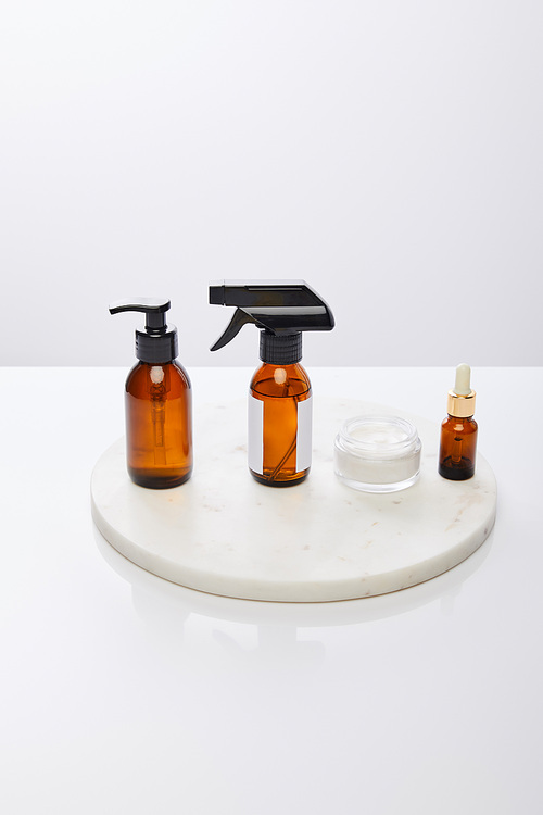 Dispenser bottles of oil next to cosmetic cream on round stand isolated on grey