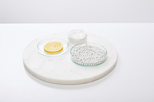 Laboratory glassware with decorative beads, slice of lemon with cosmetic cream on round stand isolated on grey