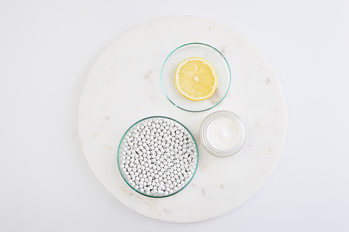 Top view of laboratory glassware with decorative beads, slice of lemon next to cosmetic cream on round stand on white background