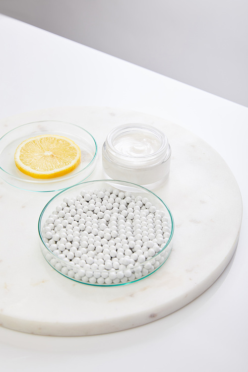 Laboratory glassware with decorative beads and slice of lemon next to cosmetic cream on round stand isolated on grey
