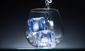 fresh vodka pouring into transparent glass with ice cubes in dark with blue back light