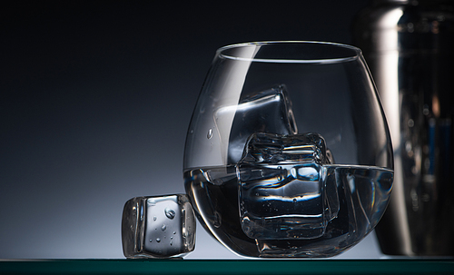 selective focus of transparent glass with ice cubes and vodka in dark with back light and shaker