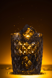 faceted glass with ice cube and splashing vodka in dark with warm back light