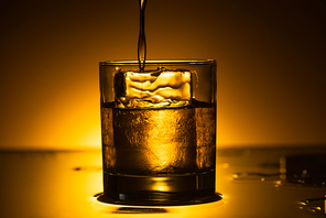 vodka pouring into transparent glass with ice cube in dark with warm back light