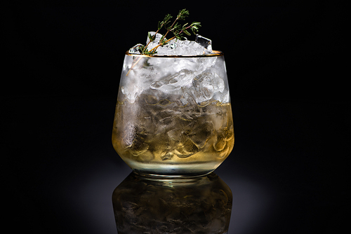 transparent glass with ice and golden liquid garnished with herb on black background