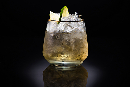 transparent glass with ice and golden liquid garnished with lime on black background