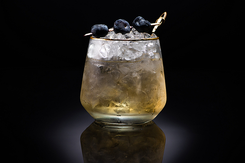 transparent glass with ice and golden liquid garnished with blueberries on black background