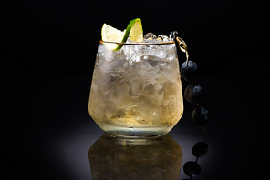 transparent glass with ice and golden liquid garnished with lime and blueberries on black background