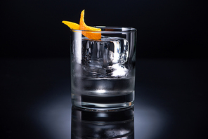transparent glass with ice cube and vodka garnished with orange peel on black background
