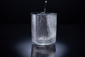faceted glass with ice and pouring vodka on black background