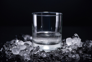 transparent glass with vodka near smashed ice on black background