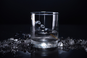 transparent glass with vodka and blueberries near smashed ice on black background
