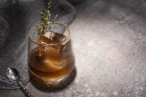 transparent glass with herb, ice cube and whiskey on concrete surface with mesh cloth and spoon