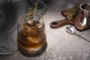 selective focus of transparent glass with herb, ice cube and whiskey on concrete surface with bar equipment